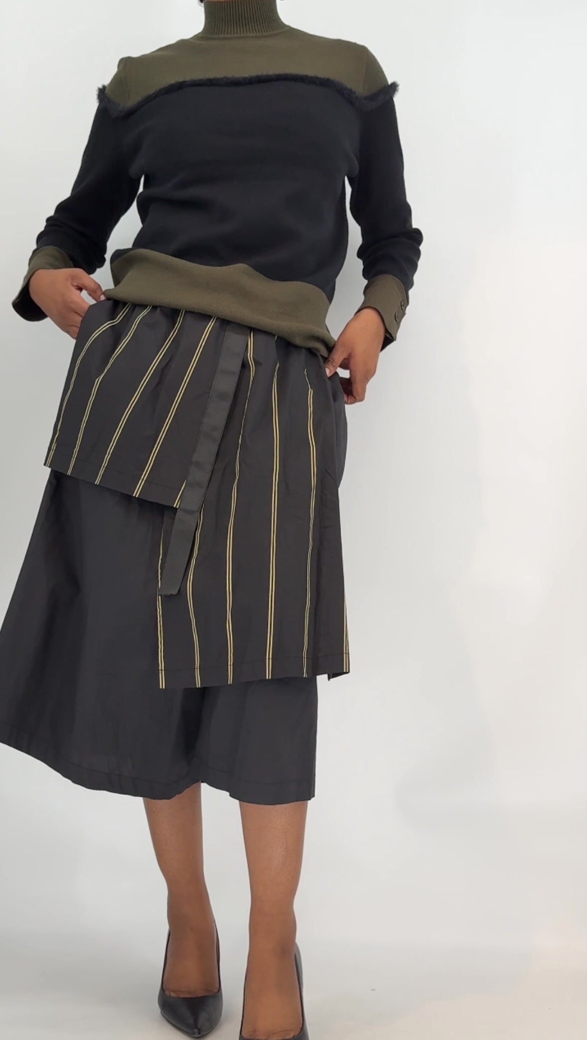Person wearing a black and green outfit with a pleated pants with layers on top on a white background.